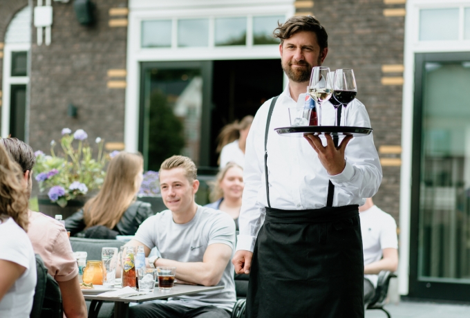 Enthousiaste horecamedewerker Pepijn serveert verschillende glazen op de juiste manier dankzij Apprendo, op de achtergrond zie je tevreden terrasgangers.